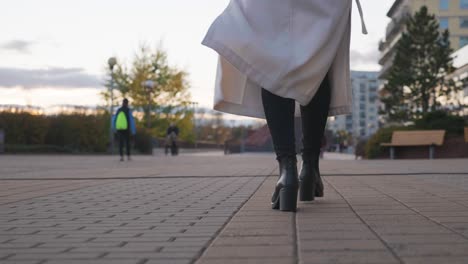 Vista-De-ángulo-Bajo-De-Una-Mujer-Caminando-Por-La-Acera-De-La-Ciudad-Y-Abrigo-Revoloteando-En-El-Viento