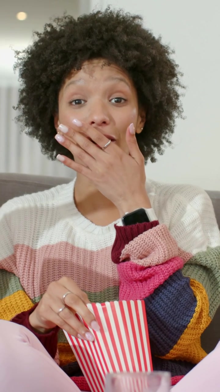 Vertical video: Laughing after friend joke, woman in striped knit reaching for popcorn tub on sofa