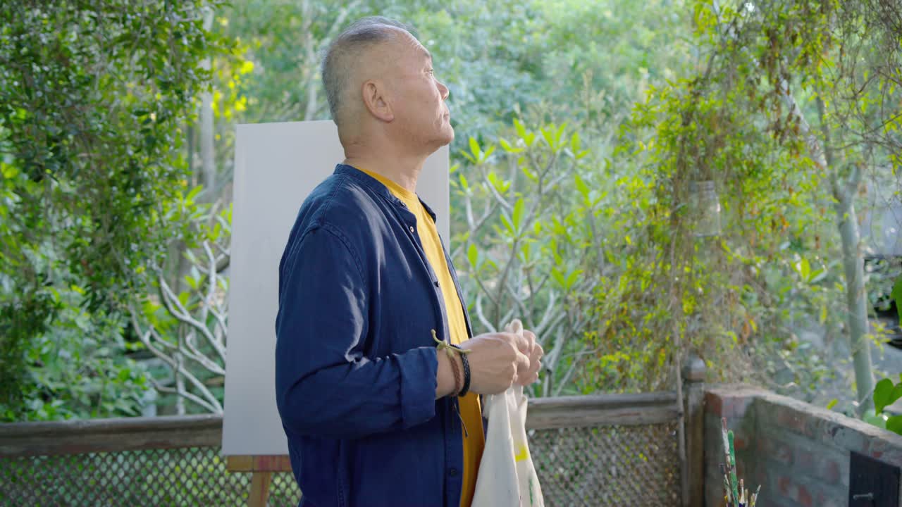 バルコニーで、アジア系の高齢男性が真っ白なキャンバスを見つめ、絵の具で汚れたエプロンを身につけ、絵を描く準備をしている。
