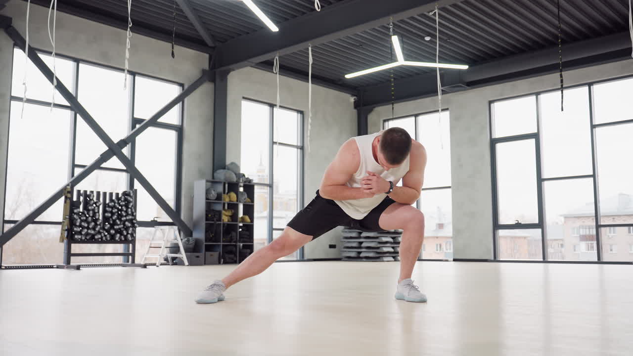 Man wearing white tank and black shorts walking heel to toe across wooden gym floor past weights rack under modern ceiling lights demonstrating balance, strength and control