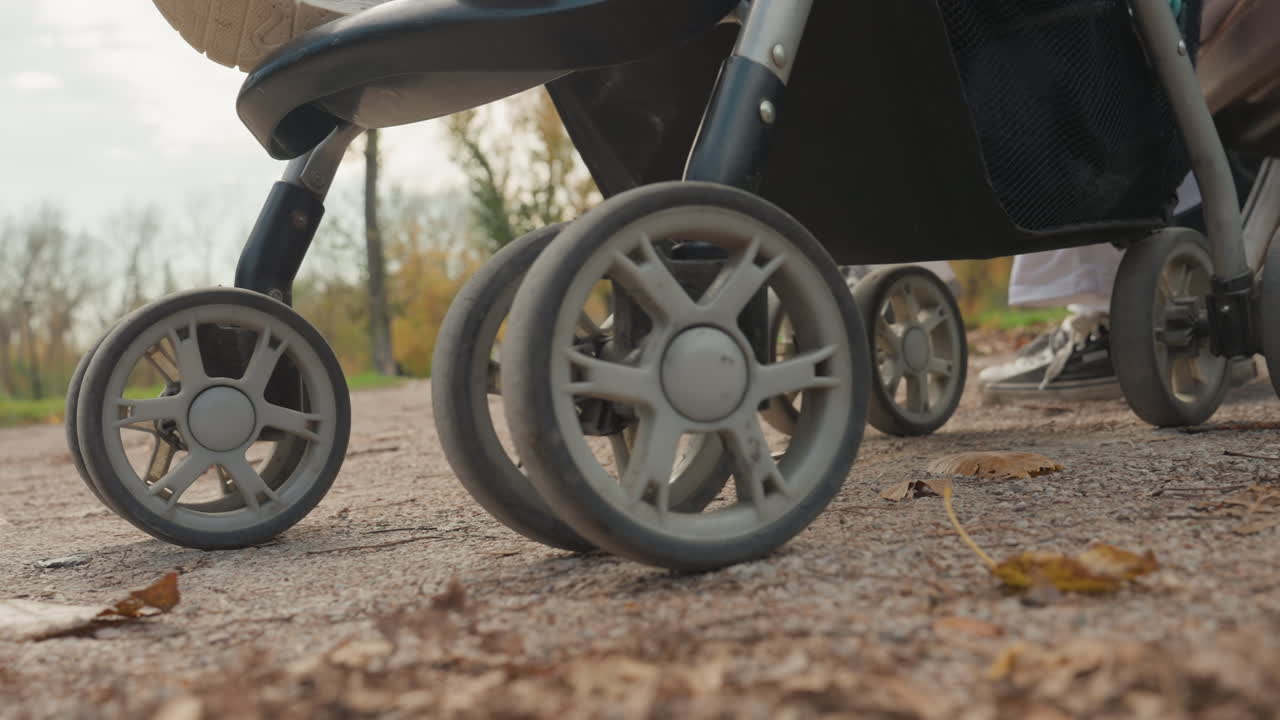 En förälder går med sitt barn utomhus. Föräldern skjuter småbarnet längs en grusgång i parken under hösten. Föräldern styr barnvagnen över gruset medan löven sprids under en pigg parkpromenad på hösten.