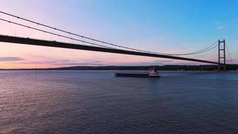 Ein-Gemächlicher-Lastkahn-Gleitet-Bei-Sonnenuntergang-Unter-Der-Humber-Bridge-Hindurch,-Festgehalten-In-Einem-Faszinierenden-Luftdrohnenclip-Während-Der-Goldenen-Stunde