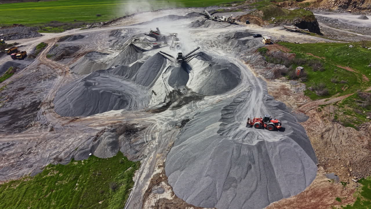 Workers operate machinery at a granite quarry during the day with piles of granite stone and dust in the air