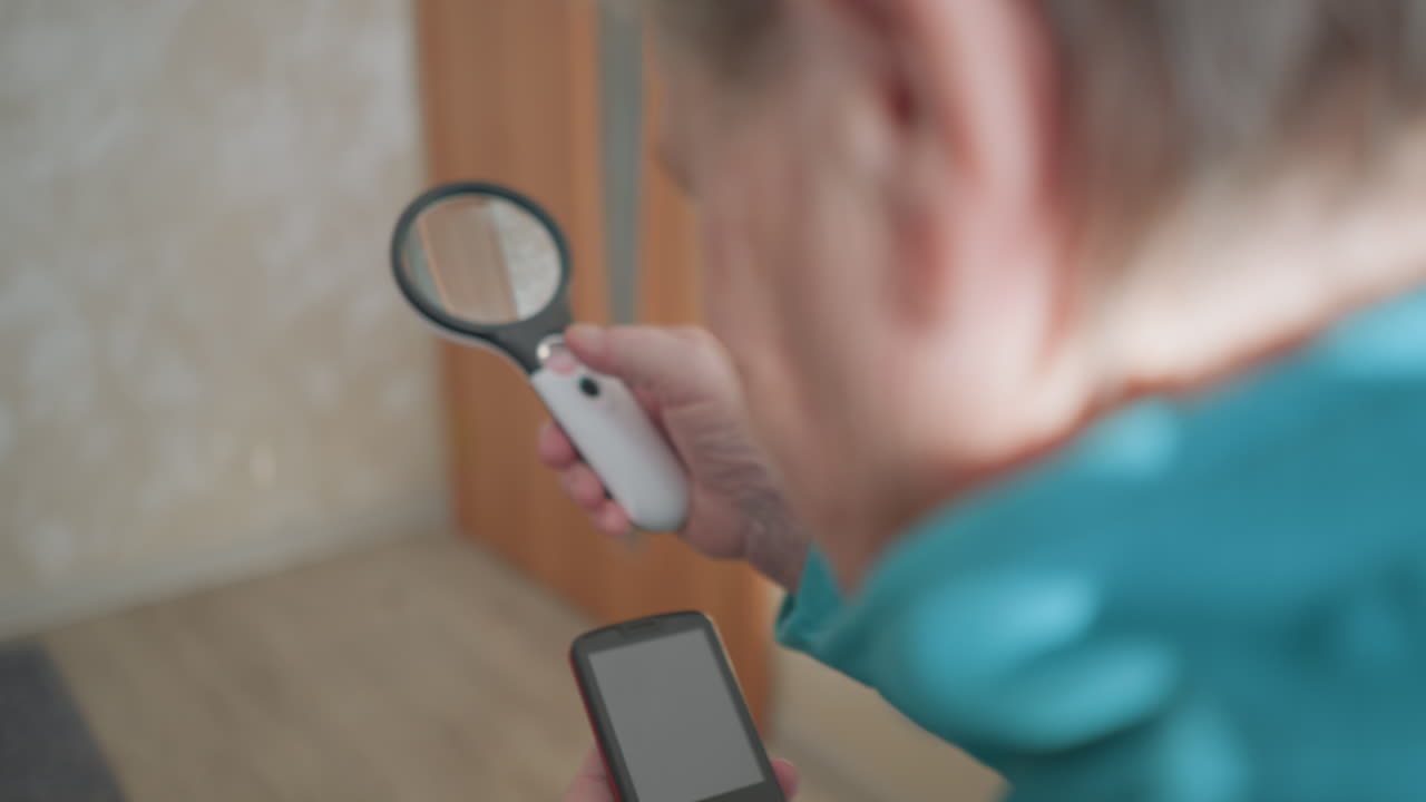 Close-up of elderly woman in green jacket holding mobile phone and magnifying glass, inspecting phone screen carefully, focused expression, well-lit indoor space, examining details on mobile device