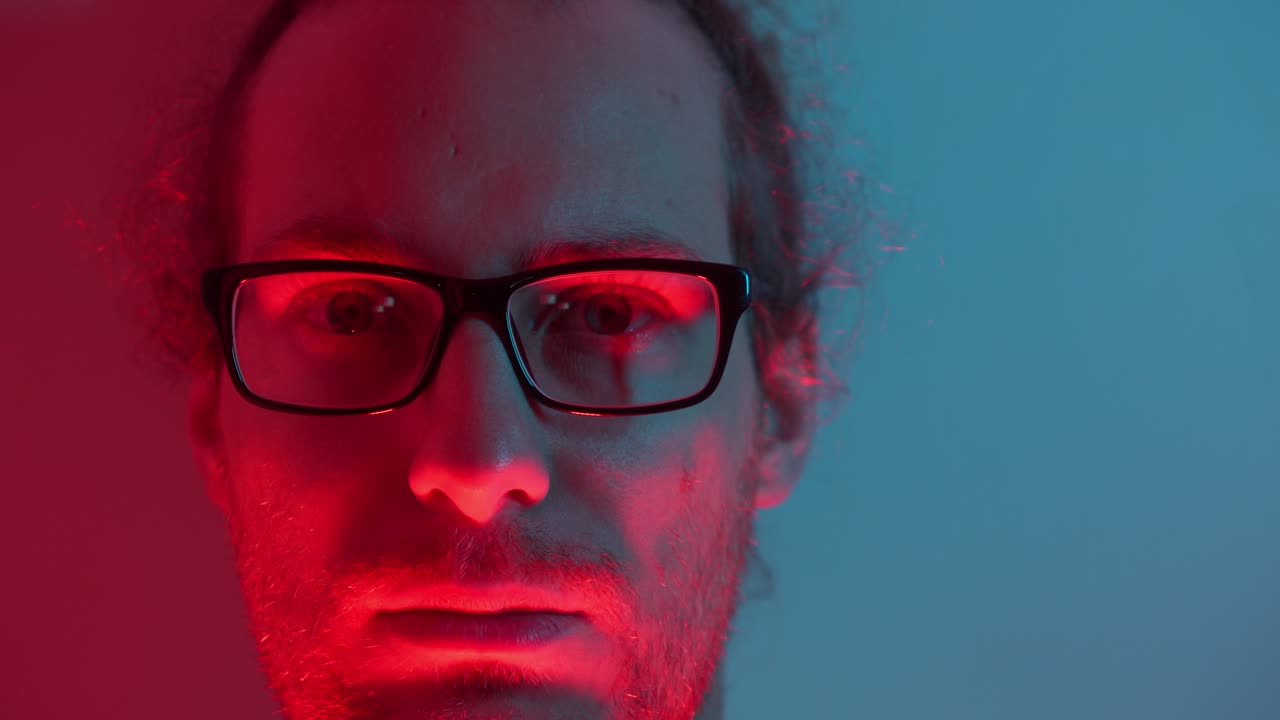 Frontal view of man with glasses in red and blue lighting, highlighting facial features, shadows dramatic as red light grows and then fades