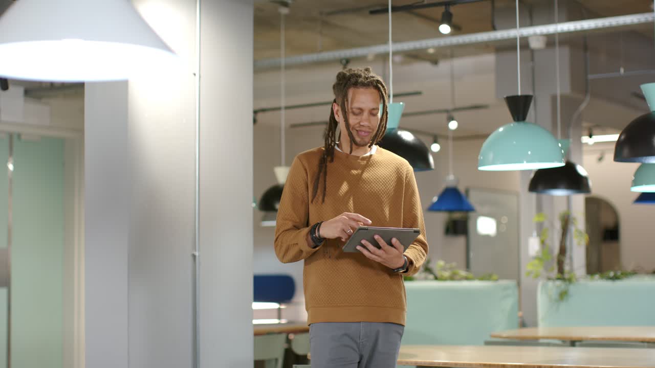 Hombre afroamericano con suéter mostaza caminando en un área de coworking, tocando una tablet, deteniéndose y sonriendo.