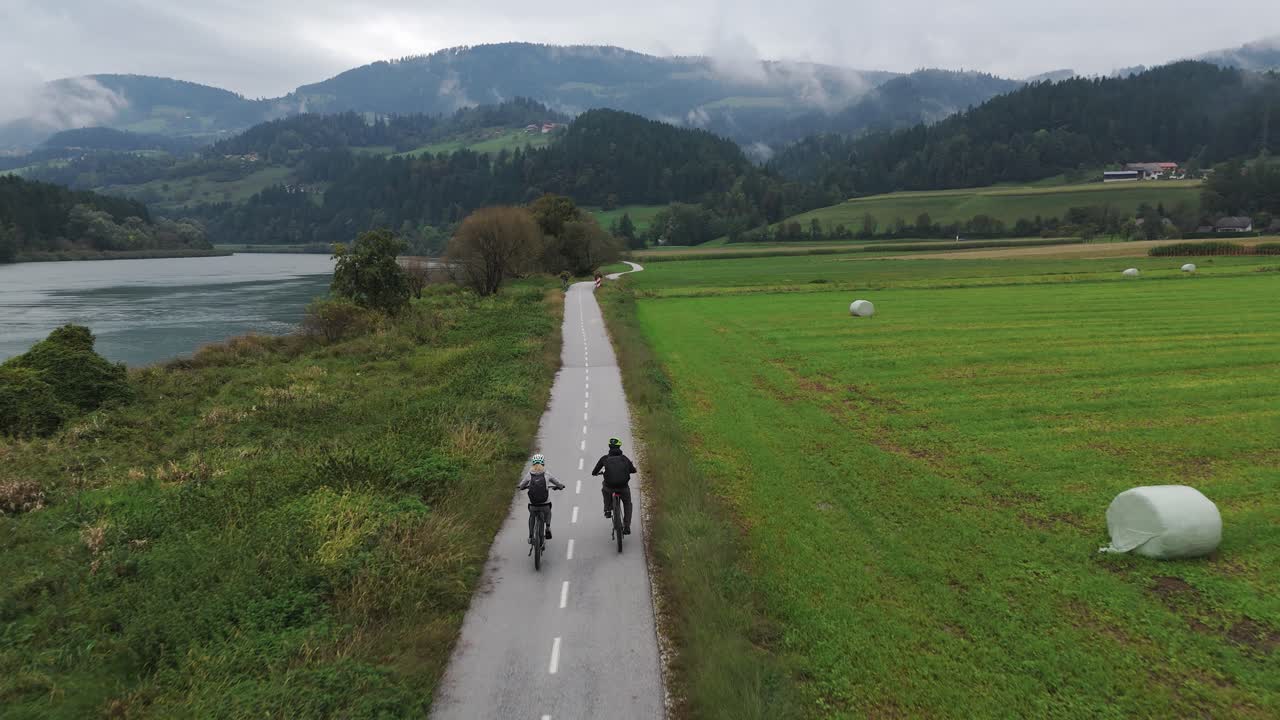 A couple rides electric bikes along a scenic riverside path through peaceful countryside with fields, forest, and distant hills on a cloudy day. Captured with smooth aerial tracking motion