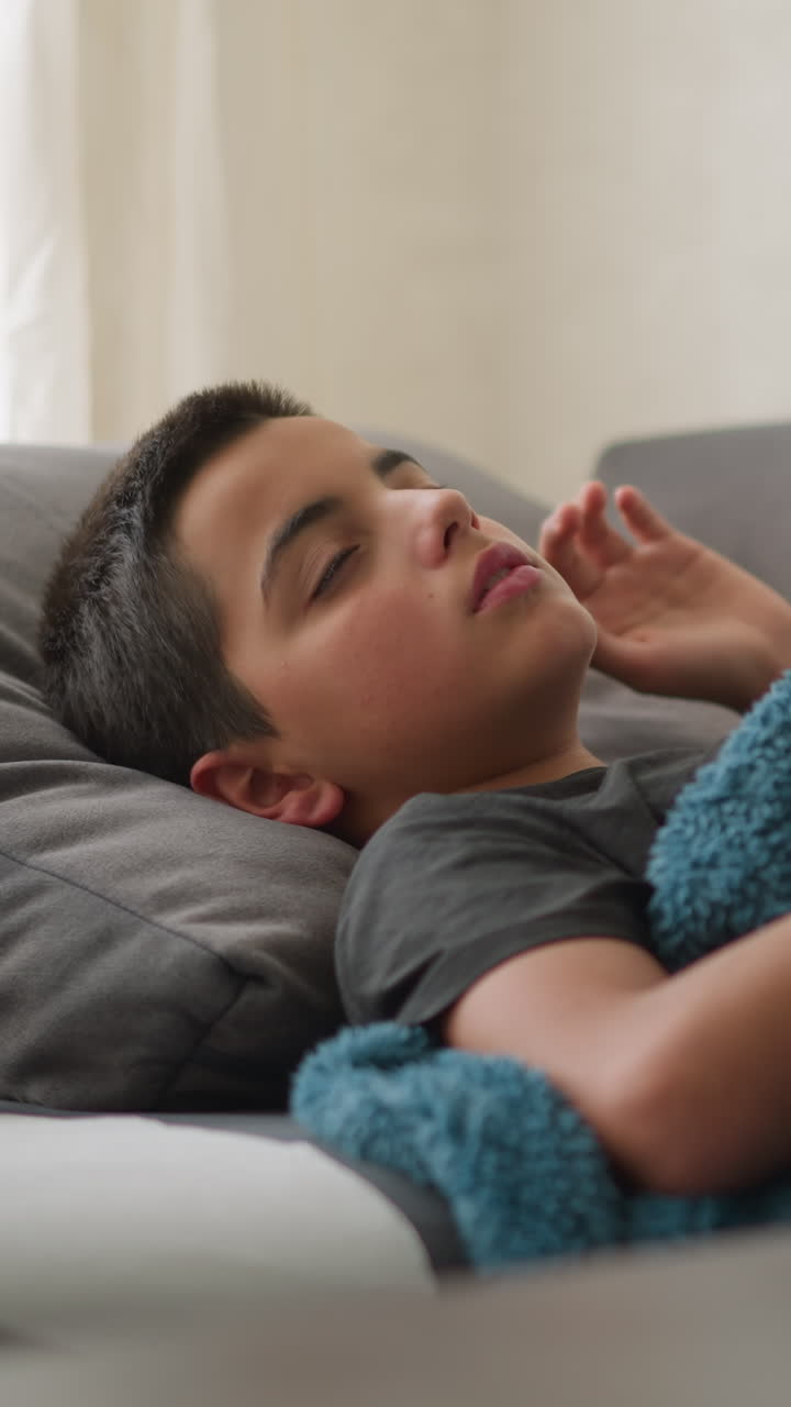 Anak laki-laki sakit menyesuaikan posisi di sofa dengan selimut, tisu bekas di dekat kepalanya, gelas cangkir di atas meja, dan tisu di permukaan dalam tampilan buram, anak beristirahat di rumah saat sakit