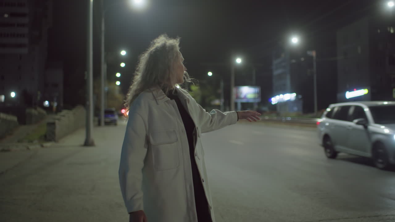 Woman in long coat stands by roadside on cold night trying to hail cab as car drives by without stopping, wrapping arms in coat while looking disappointed under city streetlights