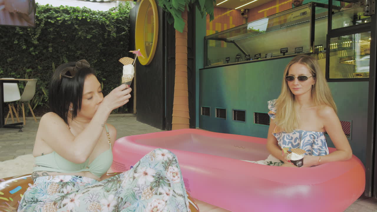 Two young women enjoy ice cream while relaxing in an inflatable pool in front of a modern ice cream parlor with tropical decor.
