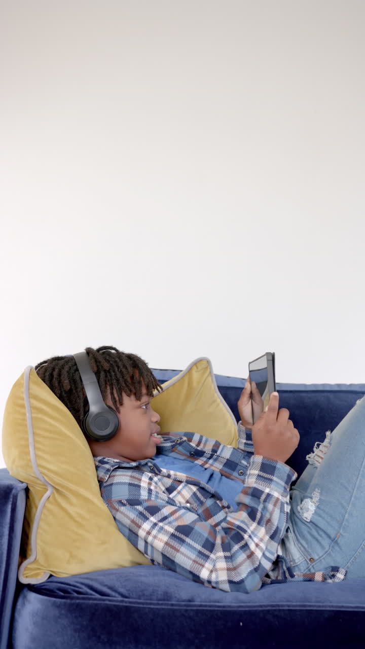 Vertical video of african american boy lying on sofa using smarphone at home, slow motion