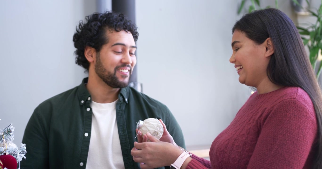 African American couple handling ornaments at home, woman in maroon knit handing bauble decorating