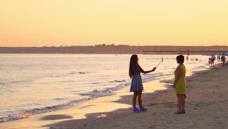 attractive girl with long hair is making selfie on the sea water background at sunset. mother and beautiful daughter walking at the seaside in the evening.