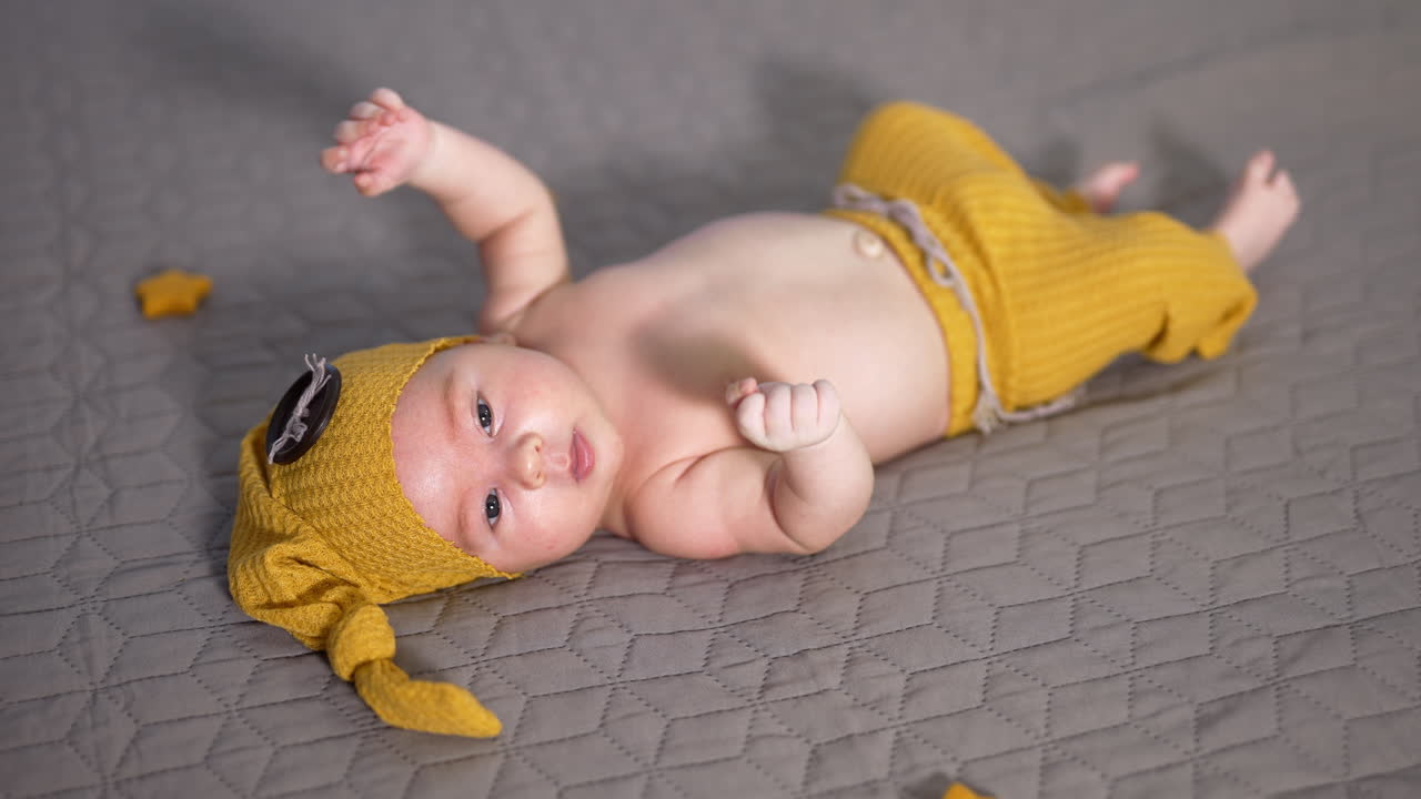 Toddler in a yellow cap with button on the bed. Baby in a funny costume tossing arms and feet. Nice kid on a grey background.