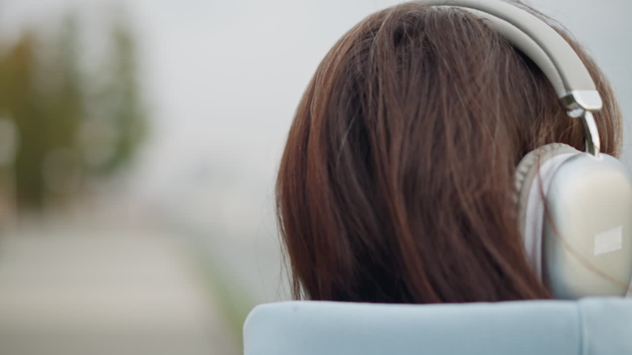 Closeup head view of lady wearing headphones, showcasing her hair and the headset, peaceful outdoor moment, focused expression, enjoying music, urban setting with blurred background