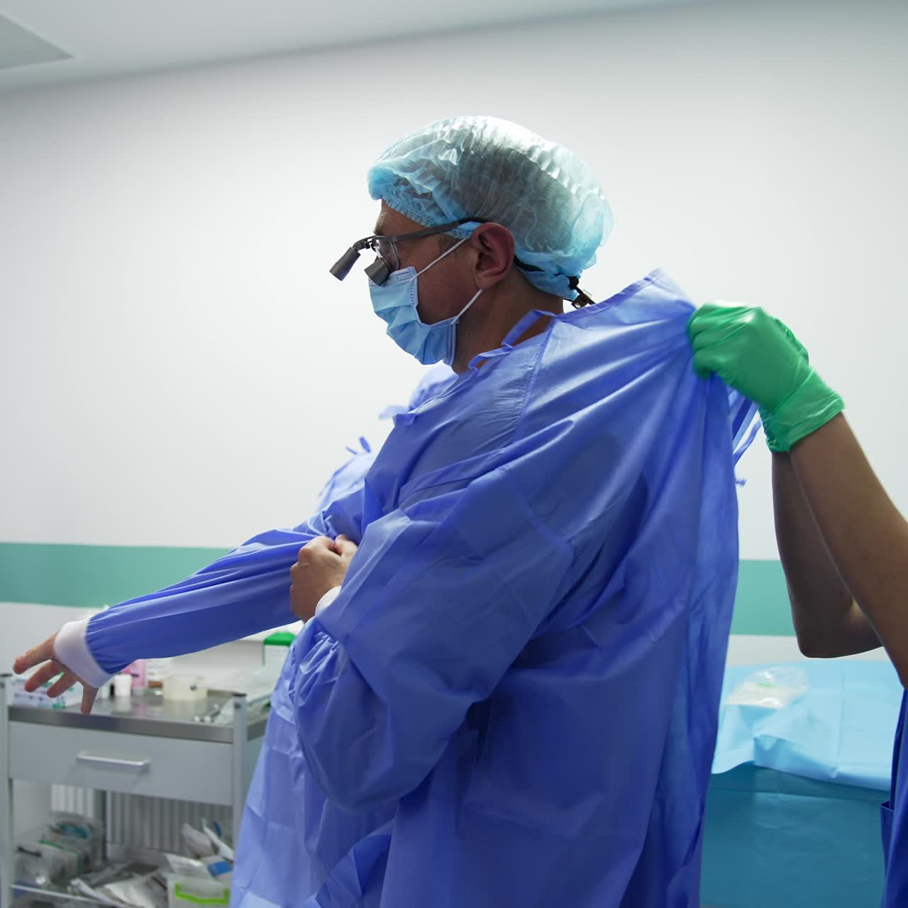 Young male assistant holds the blue robe for the surgeon. Female nurse ties the robe at the doctor's back. Surgeon getting ready for operation