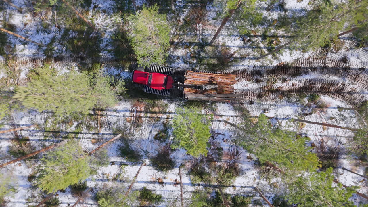 Red forwarder on snowy forest track hauling felled logs, aerial overhead riser