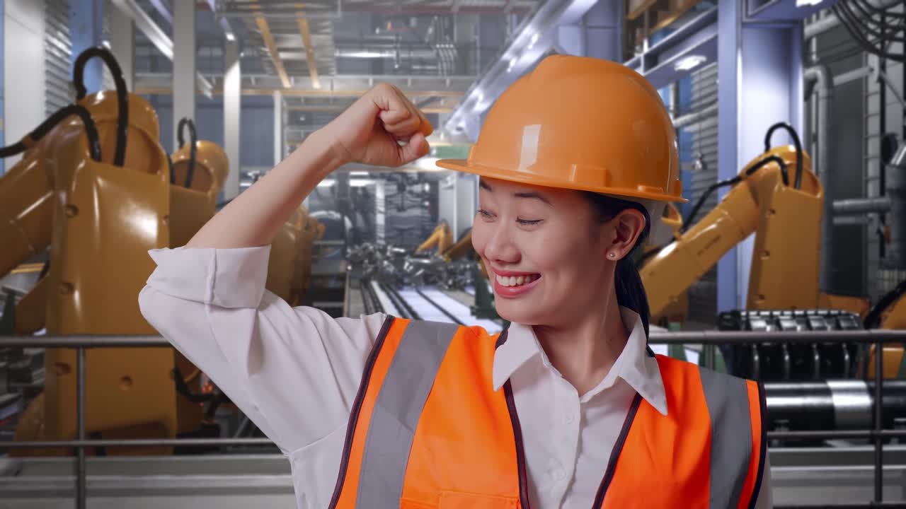 Close Up Of Asian Female Engineer With Safety Helmet Flexing Her Bicep And Smiling To Camera While Standing With Industrial Robotic Arms Working