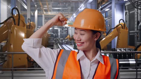 close up of asian female engineer with safety helmet flexing her bicep and smiling to camera while standing with industrial robotic arms working
