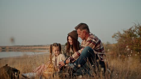 vista lateral de una familia feliz teniendo un picnic fuera de la ciudad cerca de la hierba seca y pequeños árboles verdes solitarios cerca de una orilla desierta en verano. un hombre moreno con cabello gris en una camisa a cuadros marrón vierte té para su esposa y hija pequeña de un termo durante su picnic y vacaciones fuera de la c