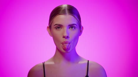 cheeky young woman sticks out tongue, purple studio light, pink background