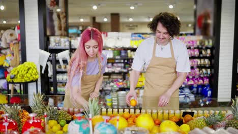 A-brunette-guy-with-curly-hair-and-his-assistant-a-girl-with-pink-hair-in-a-yellow-apron-are-sorting-through-and-taking-inventory-of-products-in-the-citrus-fruit-department-in-a-supermarket