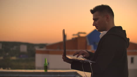un hombre con grandes auriculares blancos escribe con los dedos en el teclado de una computadora portátil de pie en el techo de un edificio al atardecer contra el fondo de la ciudad.