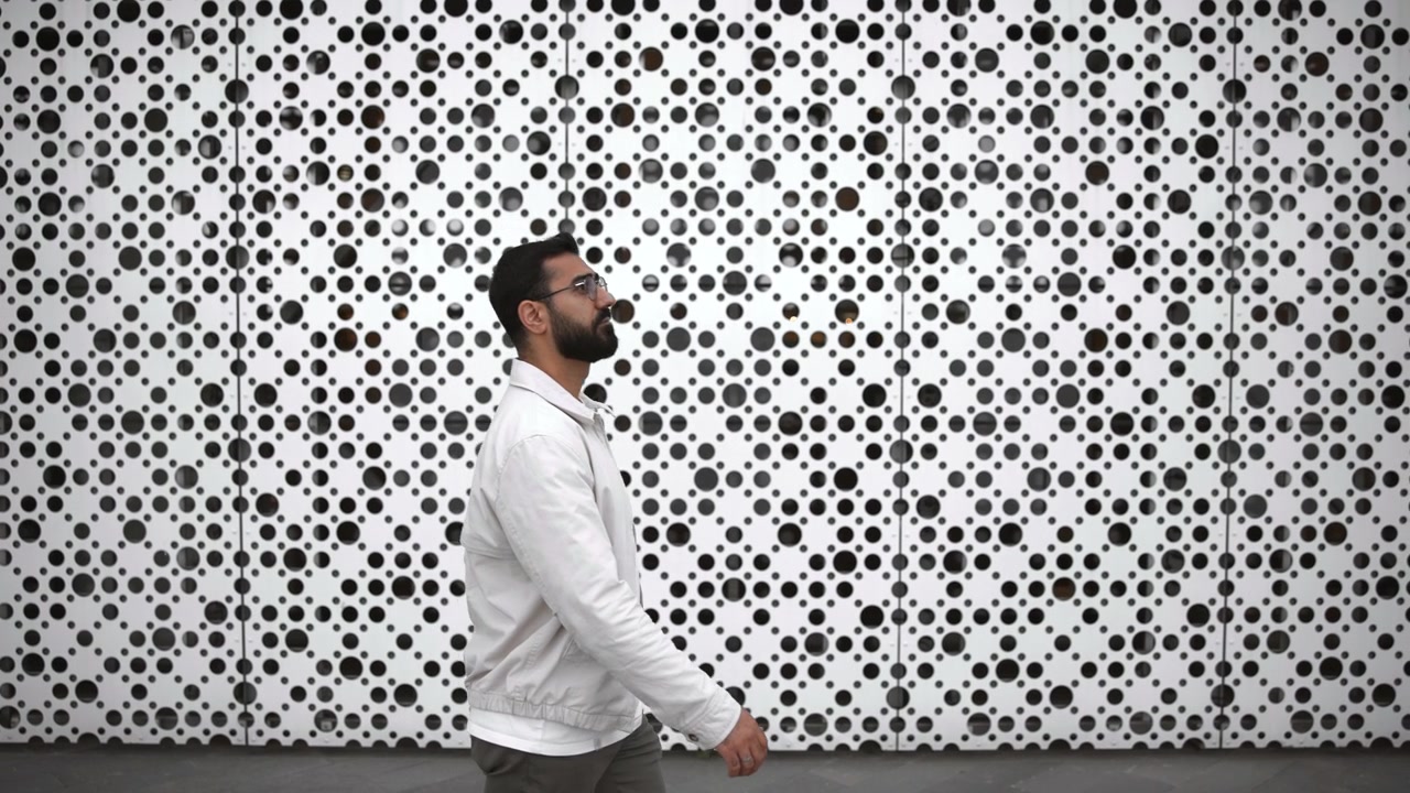 Homem adulto caminha da esquerda para a direita em primeiro plano ao lado de uma extensa parede de metal branco perfurado com recortes circulares densos, criando forte contraste, repetição, textura e arquitetura urbana moderna
