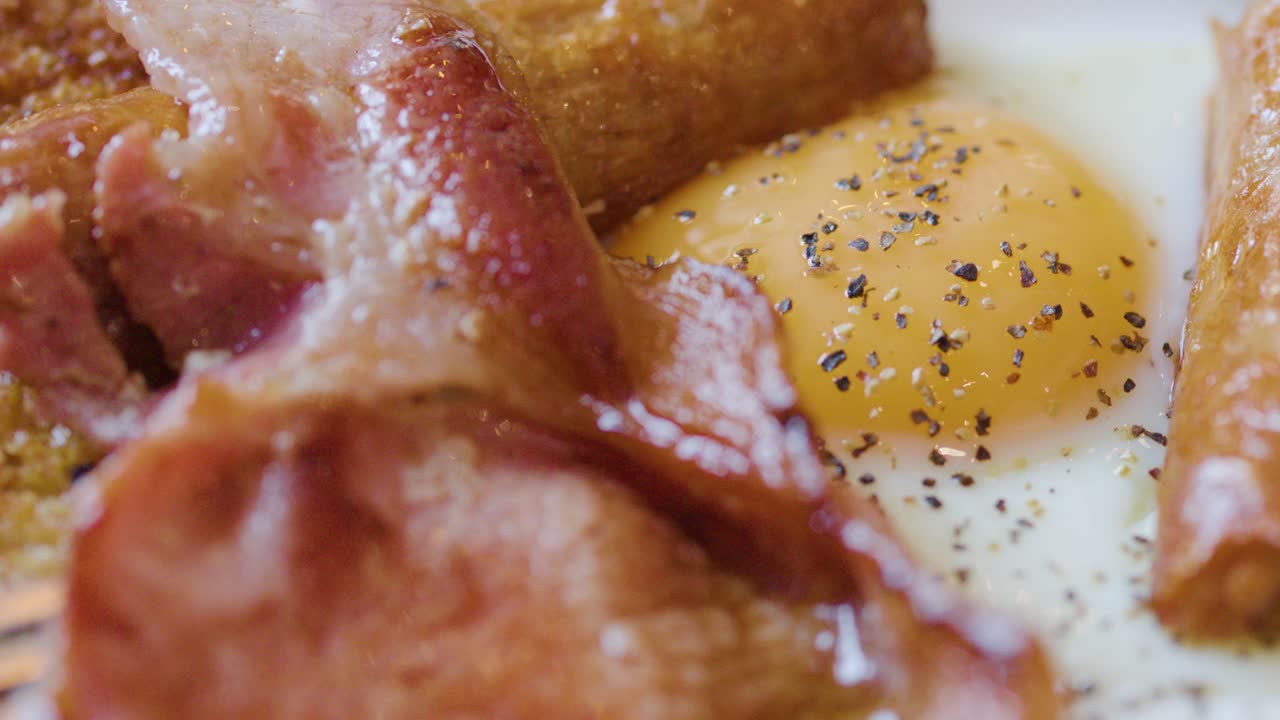 Close-up of bacon being placed beside fried egg, sausage, toast under natural morning light