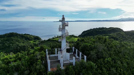 rotating aerial view of white lighthouse atop mountain in lush rainforest with vast ocean and cloud sky in background