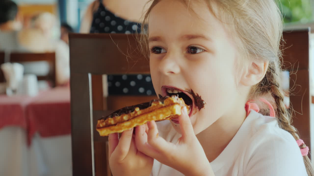 The Portrait Of A Little Girl With Two Pigtails Which Eats A Chocolate Waffle All Smeared With Sweet