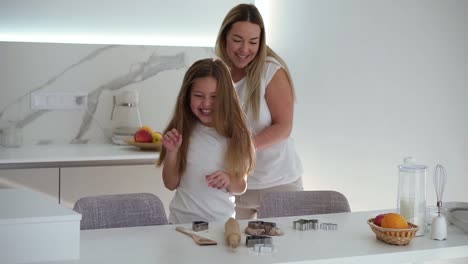 family, cooking and baking concept - happy mother and little daughter cut shaped cookies on white table at home kitchen. mom and daughter cooking and having fun together - dancing, tickling each other