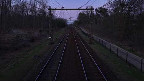 drone flies forward above dual train tracks at dusk, mimicking a train’s pov. overhead cables visible, trees and nature line both sides under soft, moody light.
