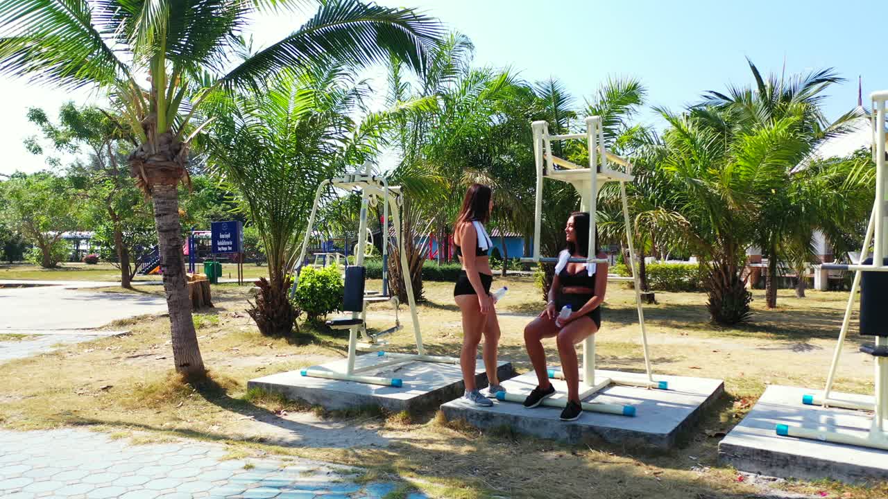 Teenage girls on outdoor fitness gym, talking about vacations on tropical island with resort near exotic beach in Thailand
