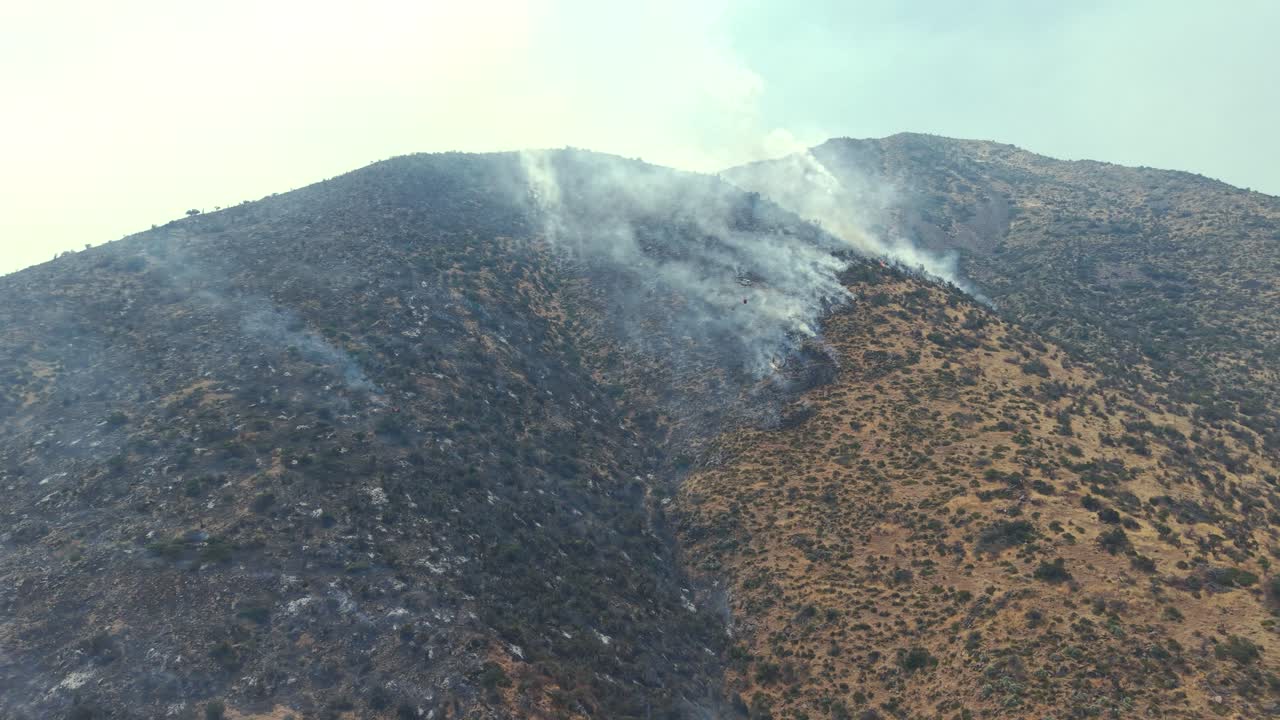 Cinematic aerial drone view of a firefighting helicopter carrying a suspended water bucket over a smoldering wildfire on a rugged mountain slope in the Andes, Chile, 4K