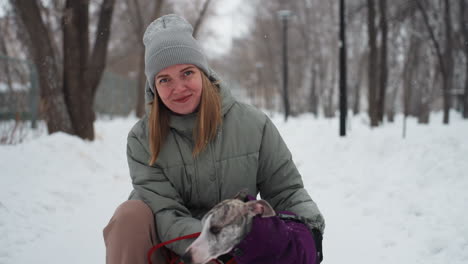 dog owner kneeling in snowy park with whippet wearing purple coat, both facing camera, woman smiling warmly, dressed in green jacket and gray beanie, peaceful winter forest scene with falling snow