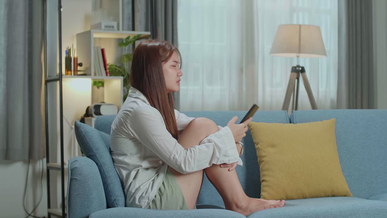 Sad Young Asian Woman Desperately Sitting On Couch And Looking At Smartphone Screen At Home