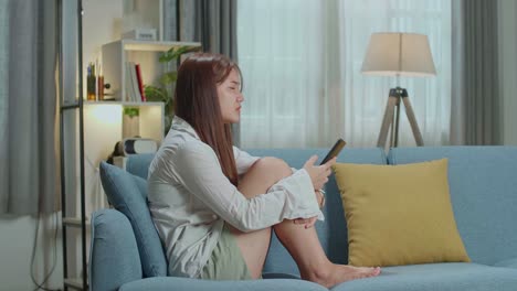 sad young asian woman desperately sitting on couch and looking at smartphone screen at home