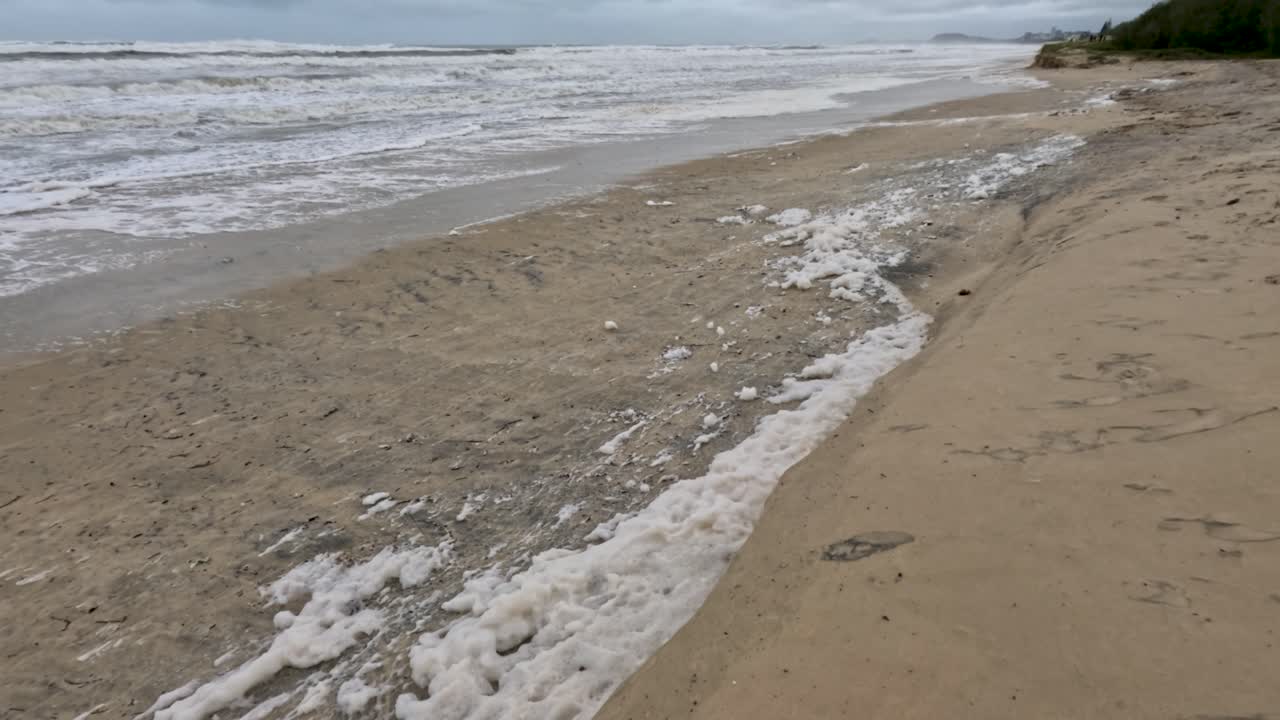 거품 모양의 파도가 모래 해변에 부히며, 구름이 가득한 하늘 아래의 사이클론 알프레드의 후유증을 보여줍니다.