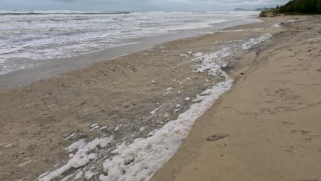 foamy waves crash onto a sandy beach, illustrating the aftermath of cyclone alfred under overcast skies