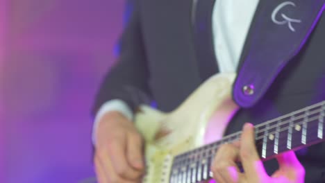 cerca de un músico profesional con traje negro tocando acordes en una guitarra eléctrica stratocaster durante una sesión en vivo en el escenario con cálidas luces de estudio en el fondo borroso