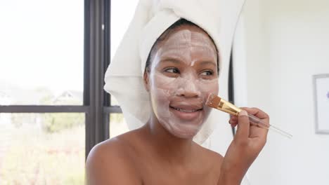 Happy-biracial-woman-with-towel-on-head-making-mask-on-face-in-bathroom,-slow-motion