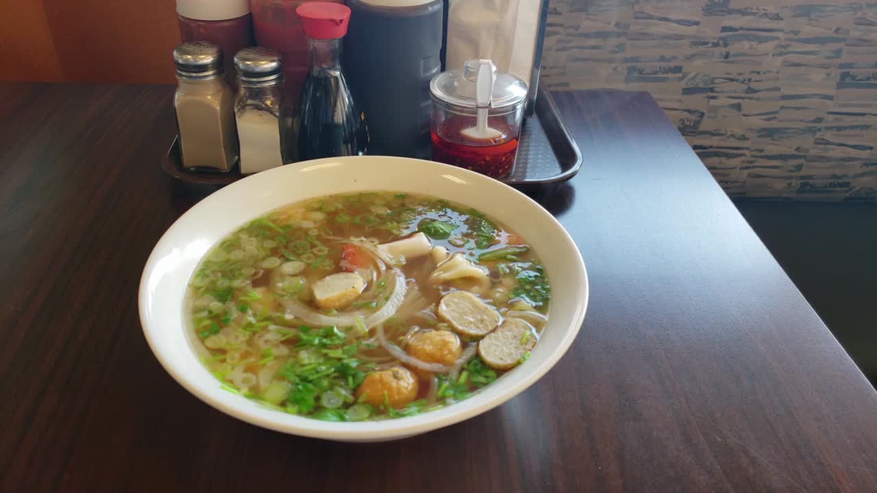 sopa de fideos pho vietnamita humeante con tubería caliente fresca fresca y humeante en un tazón junto a los condimentos tradicionales en una mesa junto a un banco en un restaurante