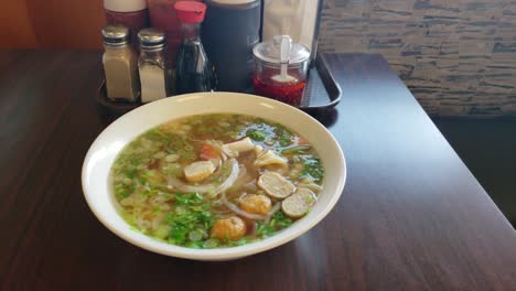 Frische-Heiße-Rohrleitungen-Dampfende-Vietnamesische-Pho-Nudelsuppe-Frisch-Und-Dampfend-In-Einer-Schüssel-Neben-Traditionellen-Gewürzen-Auf-Einem-Tisch-Neben-Einer-Bank-In-Einem-Restaurant