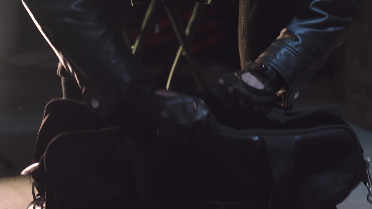 criminal con guantes de cuero abriendo un bolso negro con cremallera en el sótano