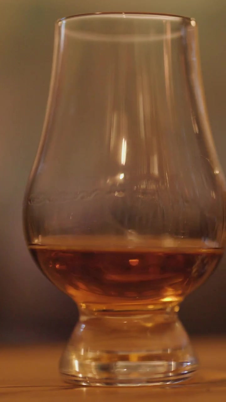 Un singolo bicchiere di whisky con liquido ambrato è posizionato su un bancone di legno, illuminato calorosamente, mentre un secondo bicchiere entra gradualmente nell'inquadratura da destra.