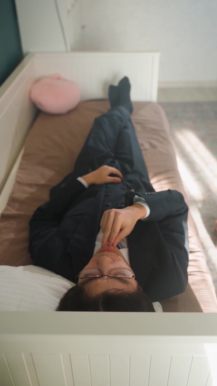 Asian businessman lying on bed, suit and glasses, wristwatch visible, hand near mouth, soft morning sunlight, pink cushion, small bedroom, quiet reflection, subtle fatigue and pause
