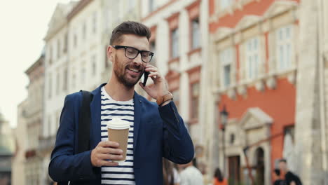 Apuesto-Hombre-De-Negocios-Sosteniendo-Una-Taza-De-Café-Para-Llevar-Y-Hablando-Por-Teléfono-Móvil-Mientras-Camina-Por-La-Calle