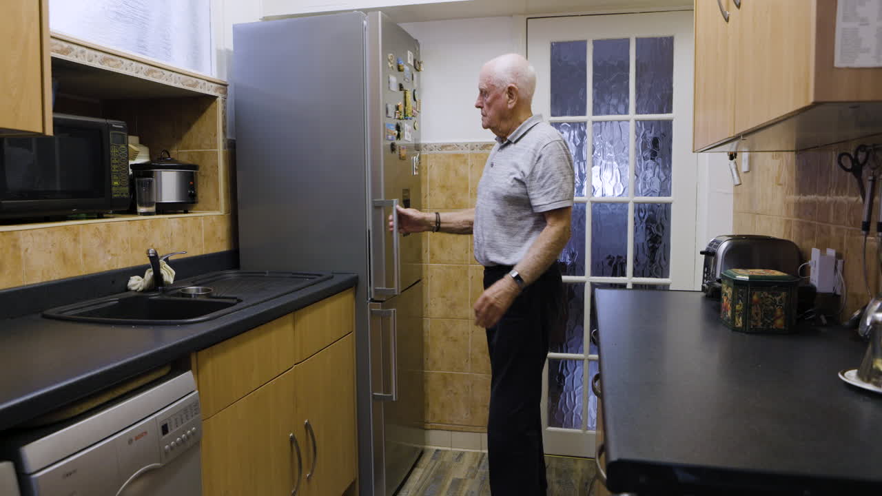 An elderly man reaches inside an open refrigerator in the kitchen