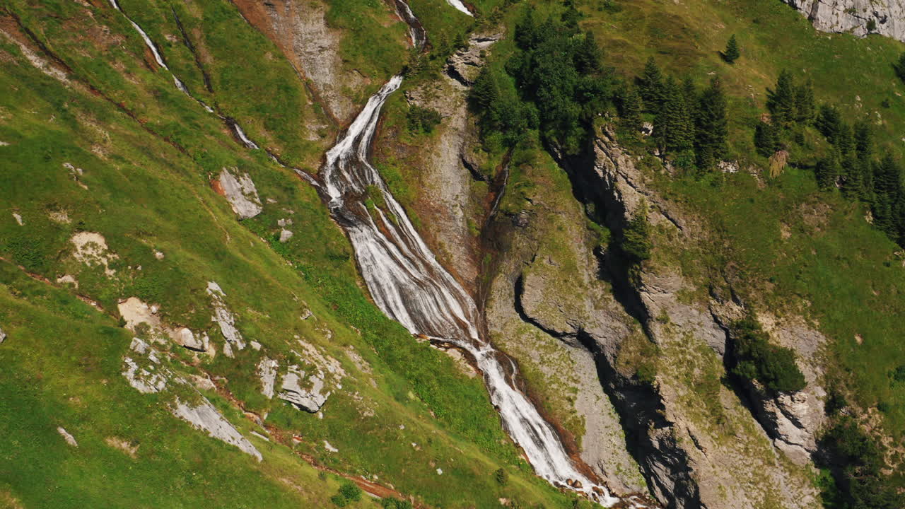 Aerial push in above alpine waterfall cascading on a steep, grassy and rocky mountainside on a sunny summer day. Orbiting drone shot above waterfall. Switzerland.
